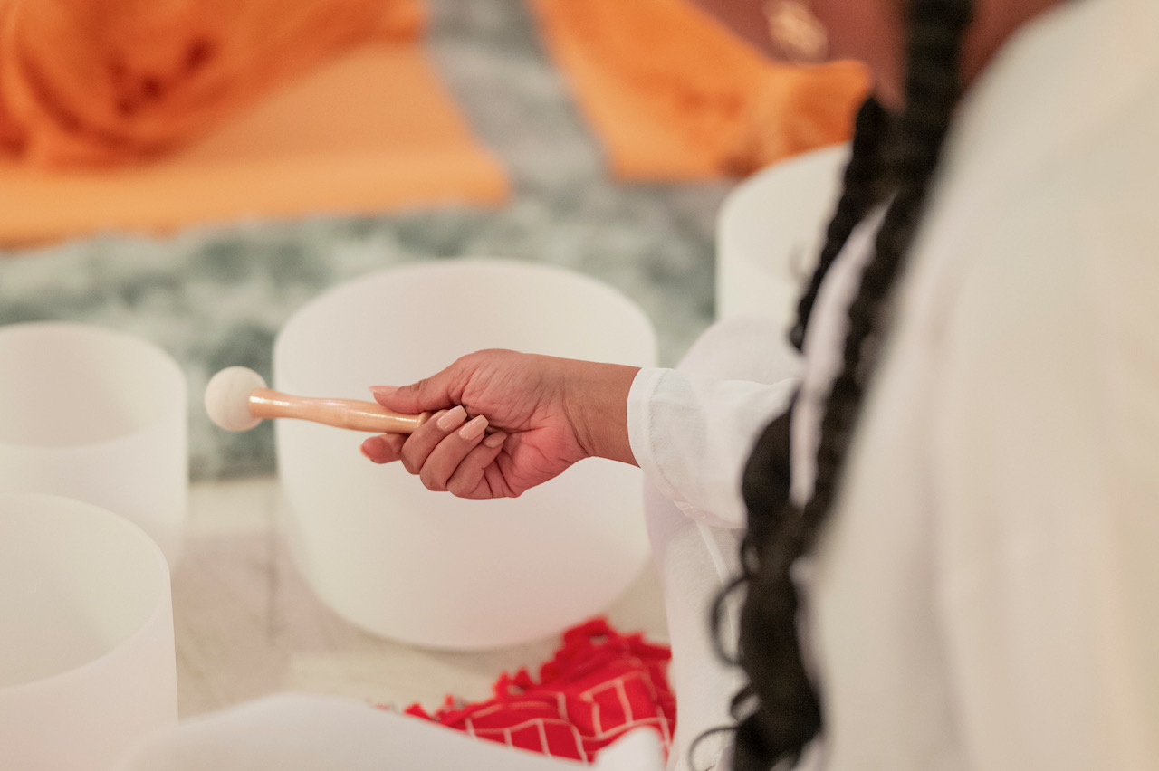 A hand poised with a mallet above a crystal singing bowl — the moment before sound becomes medicine